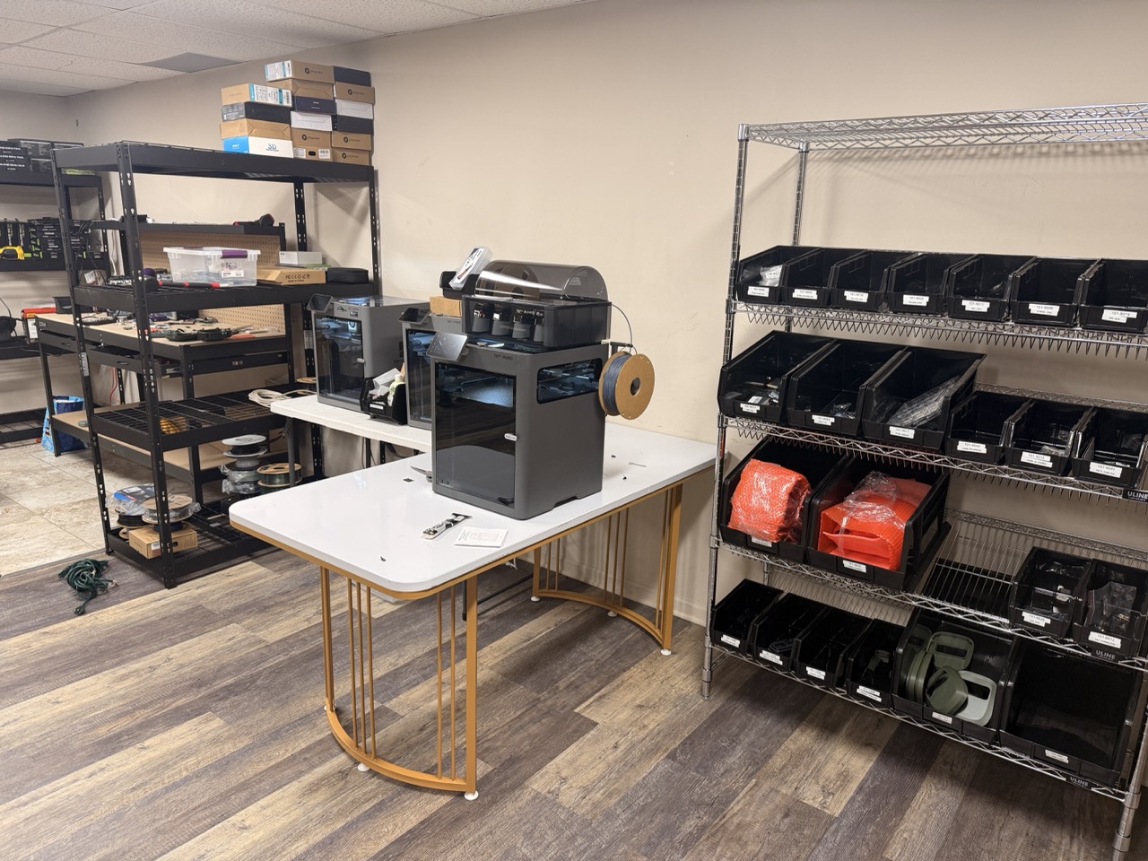 Wide view of the Waypoint Telemetry lab showing 3D printers on a work table, parts shelving, and labeled inventory bins for VisionAI One components