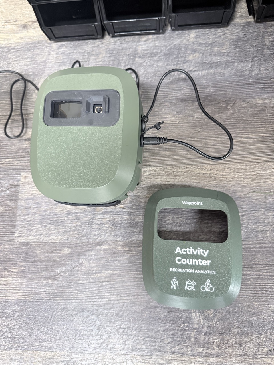 Two VisionAI One engineering samples: a green weather-sealed camera enclosure with camera window, and a labeled front bezel reading Activity Counter, Recreation Analytics, with hiker, dog, and cyclist pictograms