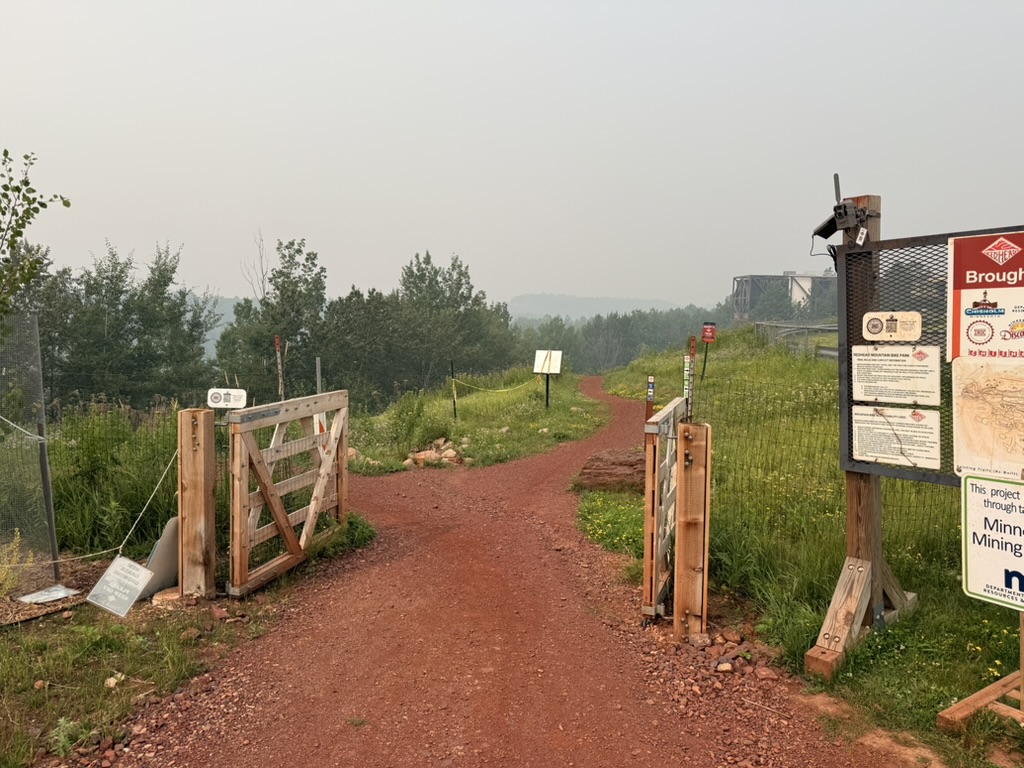 Trail camera mounted on a trailhead kiosk sign directly beside the trail gate, close and perpendicular to riders