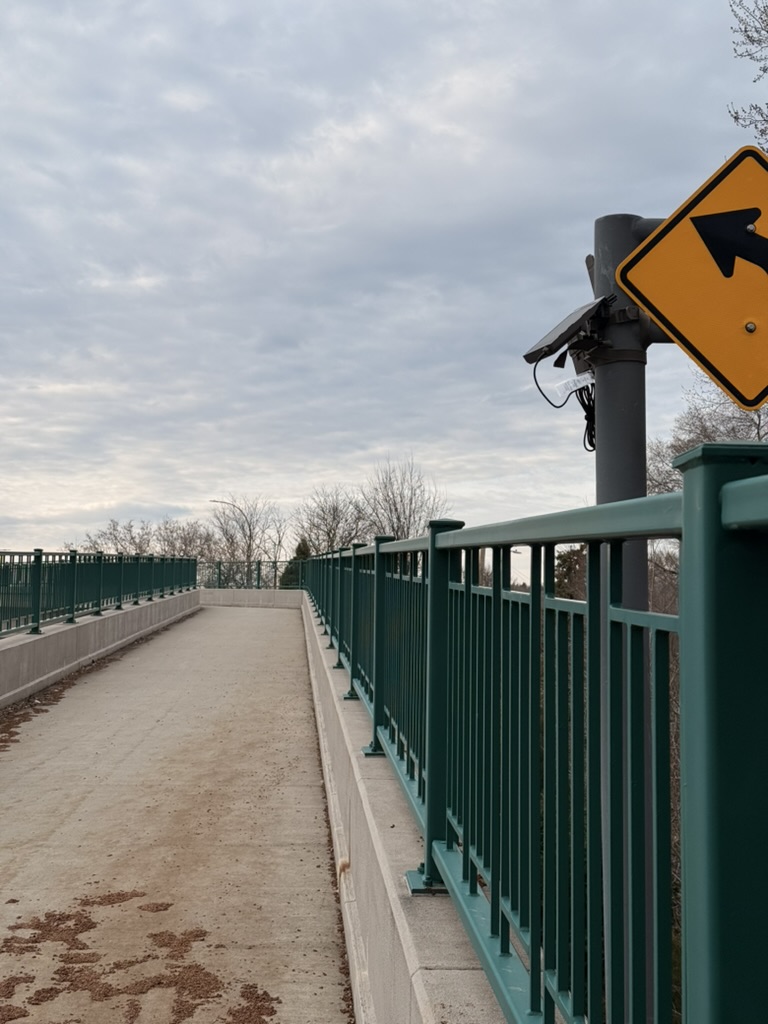 Trail camera mounted on a sign post on a pedestrian bridge, aimed down the length of the bridge deck